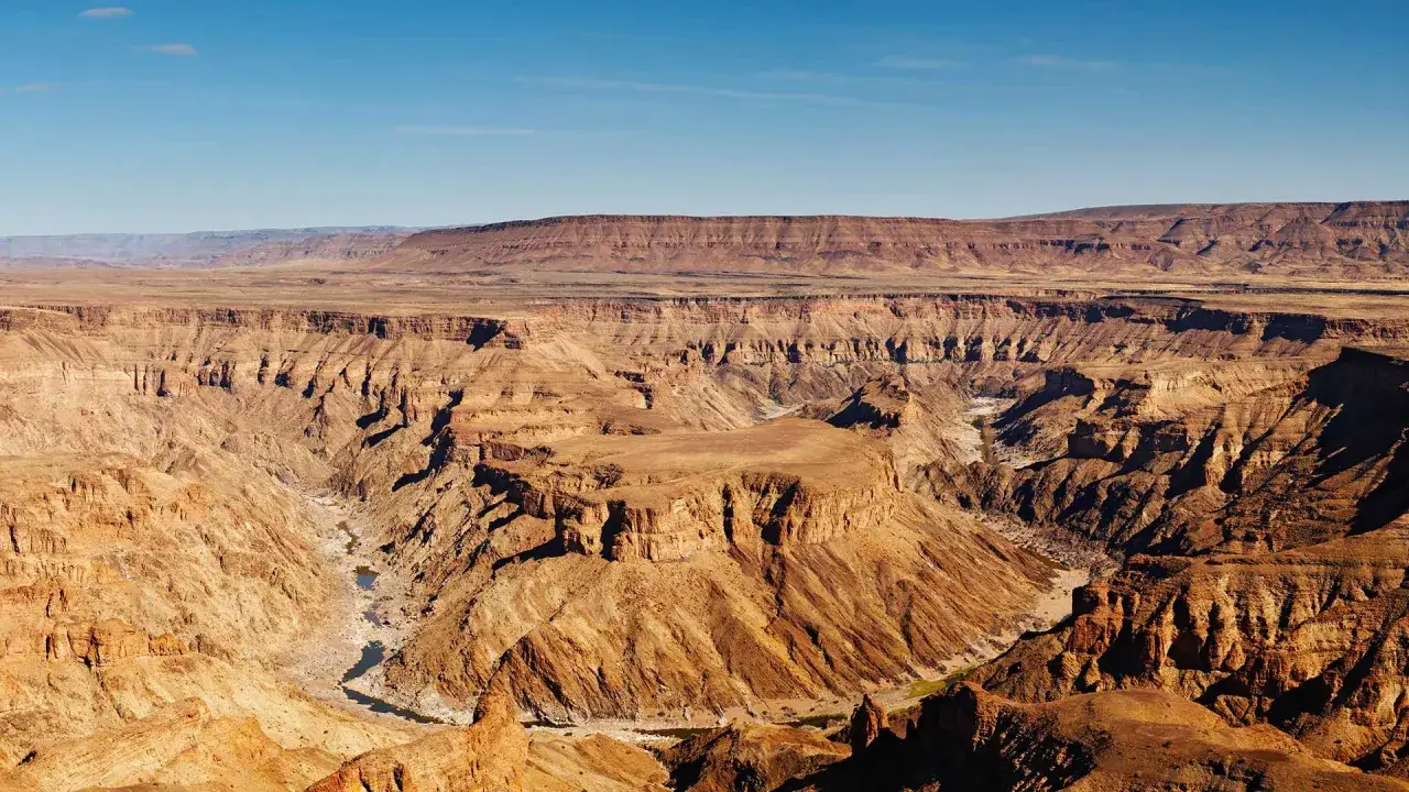 Weite, zerklüftete Namibia Landschaft mit tiefen Schluchten und einem Flussbett. Die Felsen leuchten in warmen Brauntönen unter blauem Himmel.