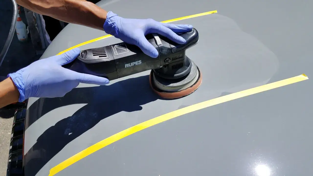 Gloved hands operate a Rupes polisher on a car's surface, marked with yellow tape. It's a bright day, raising the question: can you wax a car in the sun?