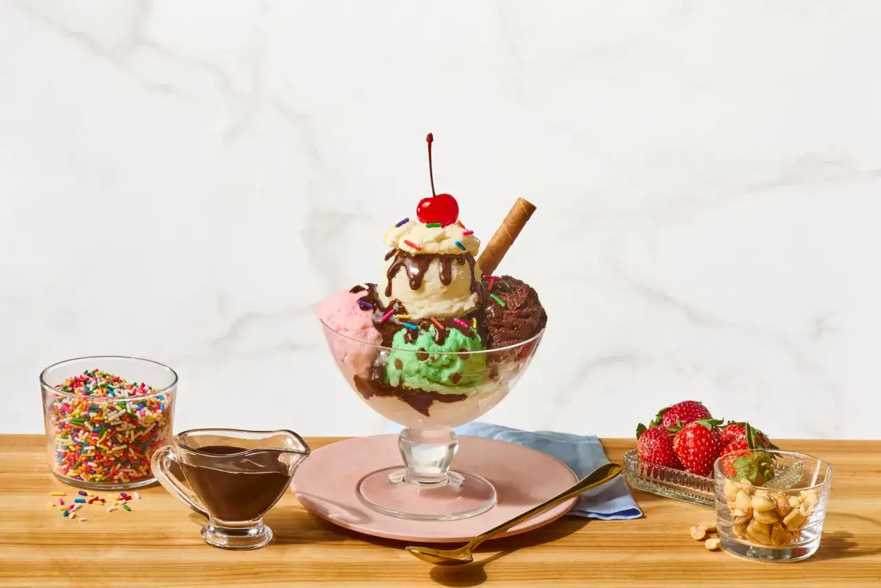 Ein Eisbecher mit drei Sorten Eis, Schokoladensauce, Streuseln, einer Kirsche und einem Waffelröllchen. Daneben Erdbeeren, Nüsse und Streusel.