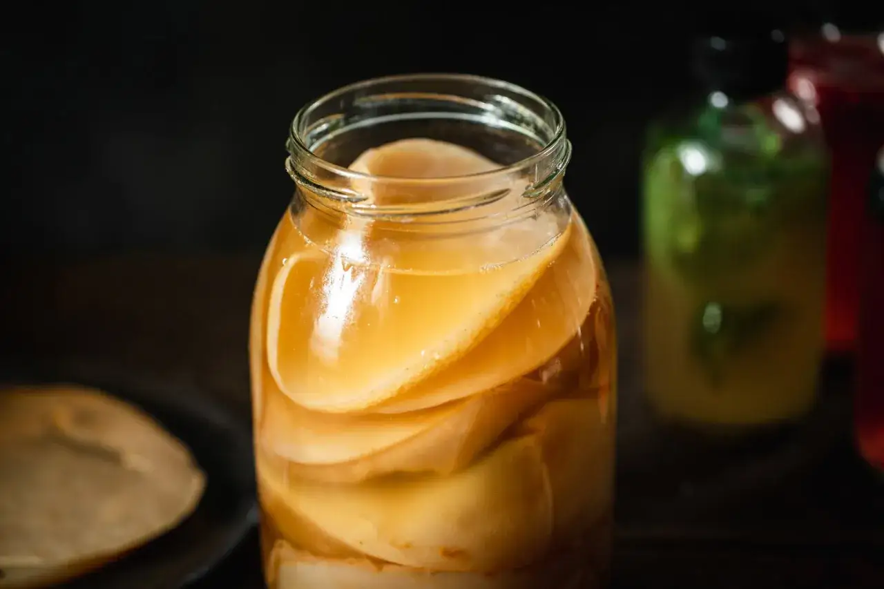 Gros plan sur un bocal rempli de couches de scoby maison, prêt pour la fermentation. Des bouteilles colorées en arrière-plan.