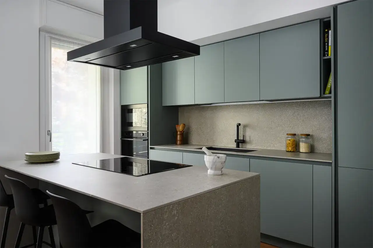 Cucina moderna con isola centrale e piano in **ardesia**. Ante color salvia, cappa nera e dettagli eleganti.
