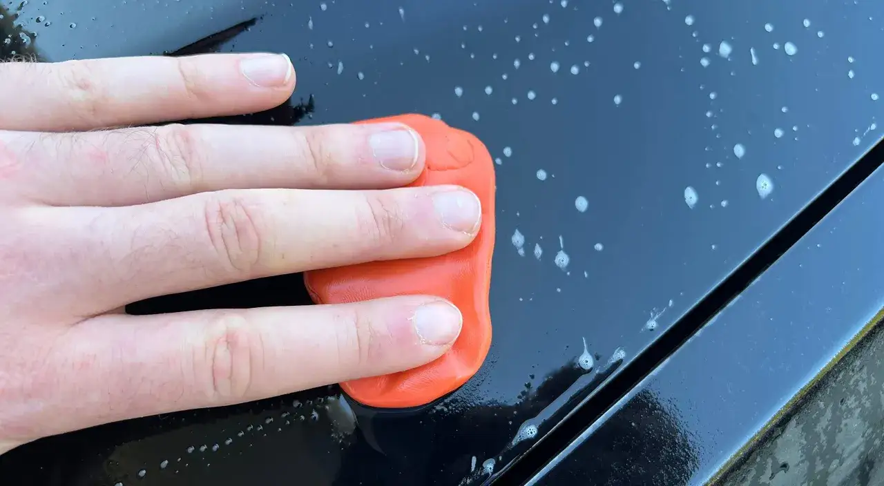 A hand glides an orange clay bar across a wet, black surface, demonstrating how a clay bar works by lifting contaminants.