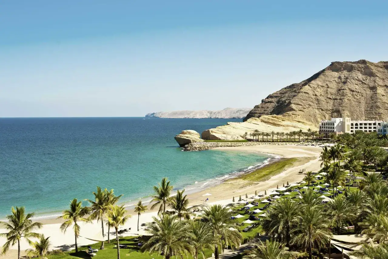 En drömresa till Oman: en vacker strand med turkost vatten, palmer och ett lyxigt hotell vid foten av berg.