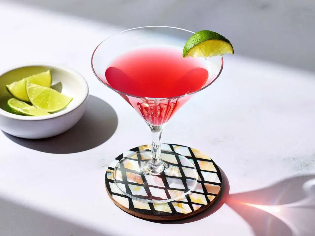 Idealny Cosmopolitan przepis: różowy drink w kieliszku, ozdobiony limonką, obok miseczka z ćwiartkami limonki.