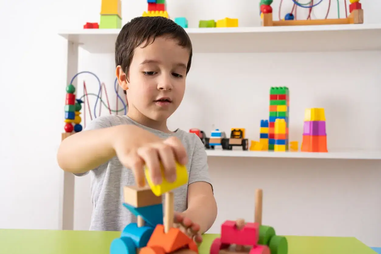 Ni&ntilde;o concentrado construyendo con bloques de madera, ideales para actividades para ni&ntilde;os de 3 a&ntilde;os. Juguetes coloridos en estanter&iacute;as.
