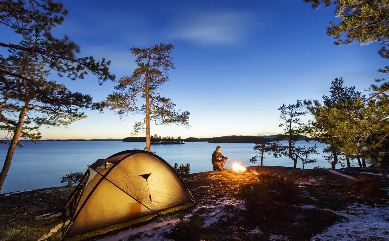 Ein Zelt und ein Lagerfeuer am Seeufer, ein Mann sitzt davor. Ein friedliches Bild von **Wildcampen Finnland** bei Dämmerung.