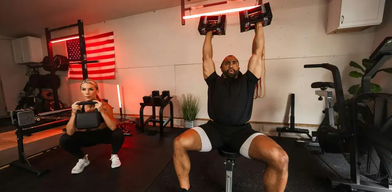 Mann hebt Kurzhanteln, Frau macht Kniebeugen. Ihr Home-Gym ist mit der besten Hantelbank und Trainingsger&auml;ten ausgestattet.
