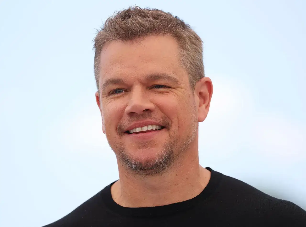 Uśmiechnięty Matt Damon, którego wiek widać po zmarszczkach wokół oczu, w czarnej koszulce na tle nieba.
