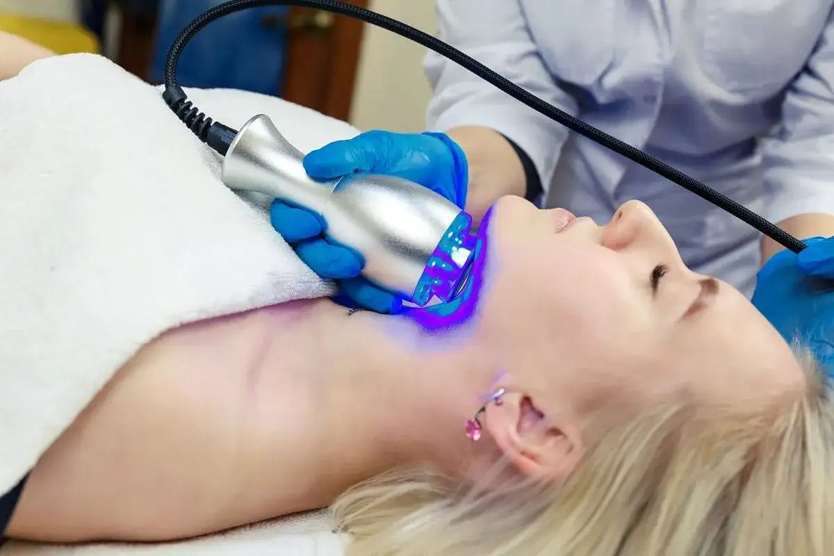 Kobieta przechodzi zabiegi dermatologiczne z użyciem urządzenia emitującego niebieskie światło na szyję.