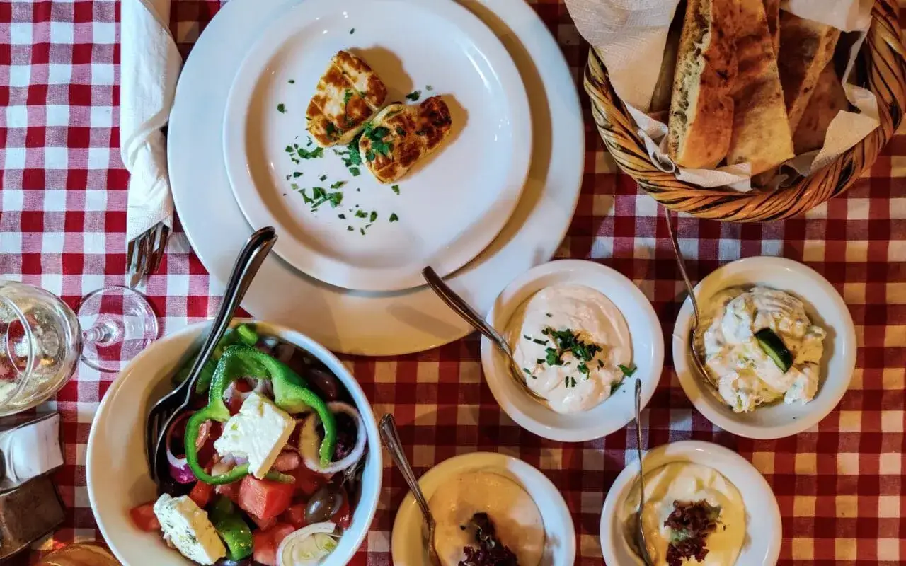 Pyszne jedzenie z Cypru: sałatka grecka, halloumi, dipy i pieczywo na czerwono-białym obrusie.