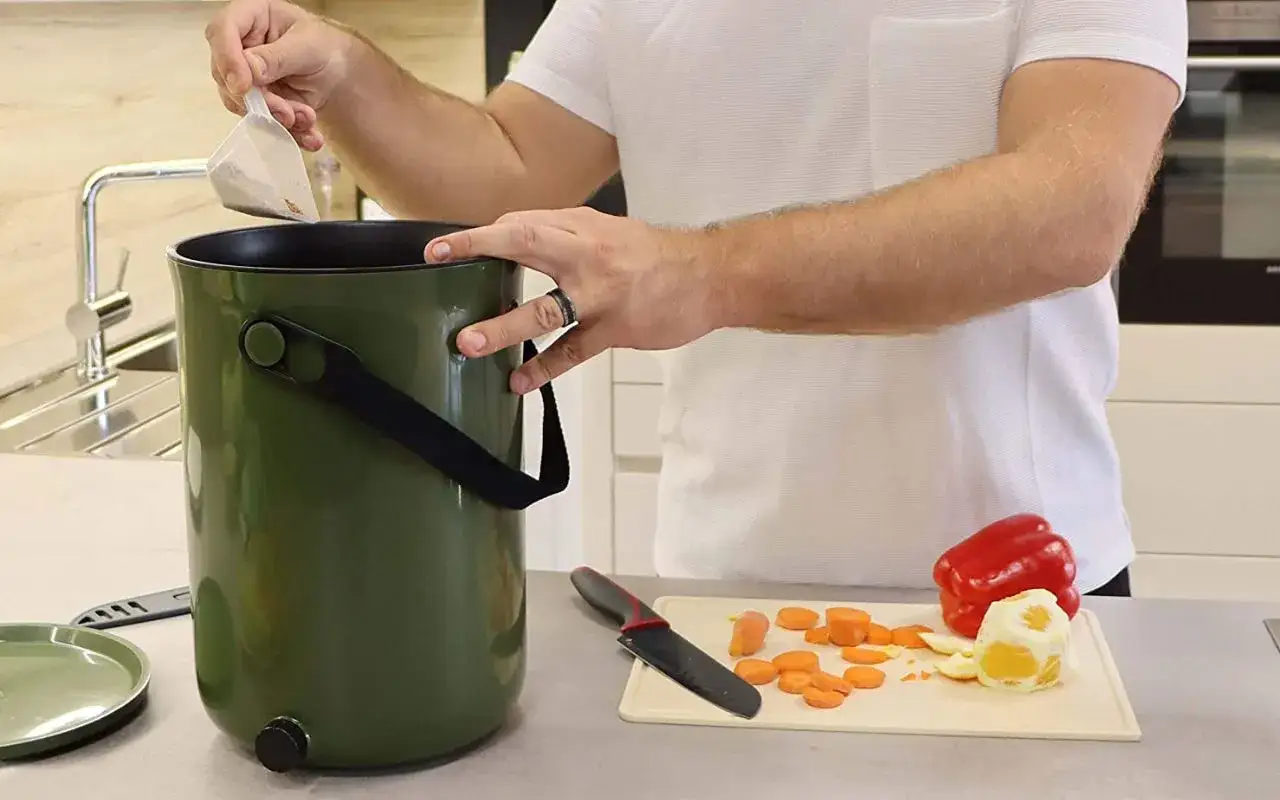 Un homme ajoute un sachet dans un seau vert, un ustensile pratique pour le compost appartement. Des carottes coup&eacute;es et un poivron rouge sont sur une planche &agrave; d&eacute;couper.