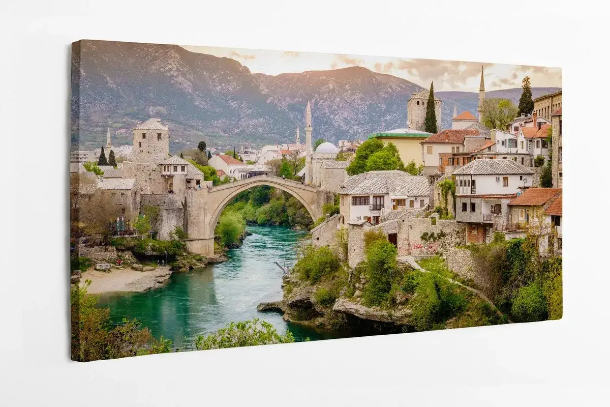 Mostar most nad turkusową rzeką, otoczony kamiennymi domami i zielenią, z górami w tle.