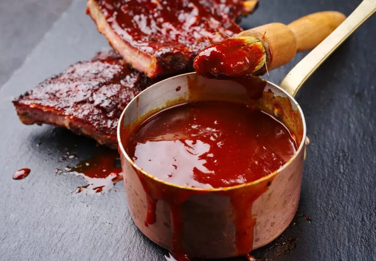 Domowy sos BBQ z wędzonym aromatem bez grilla. Pędzelek z sosem nad rondelkiem z gładkim, ciemnoczerwonym sosem i żeberkami w sosie.
