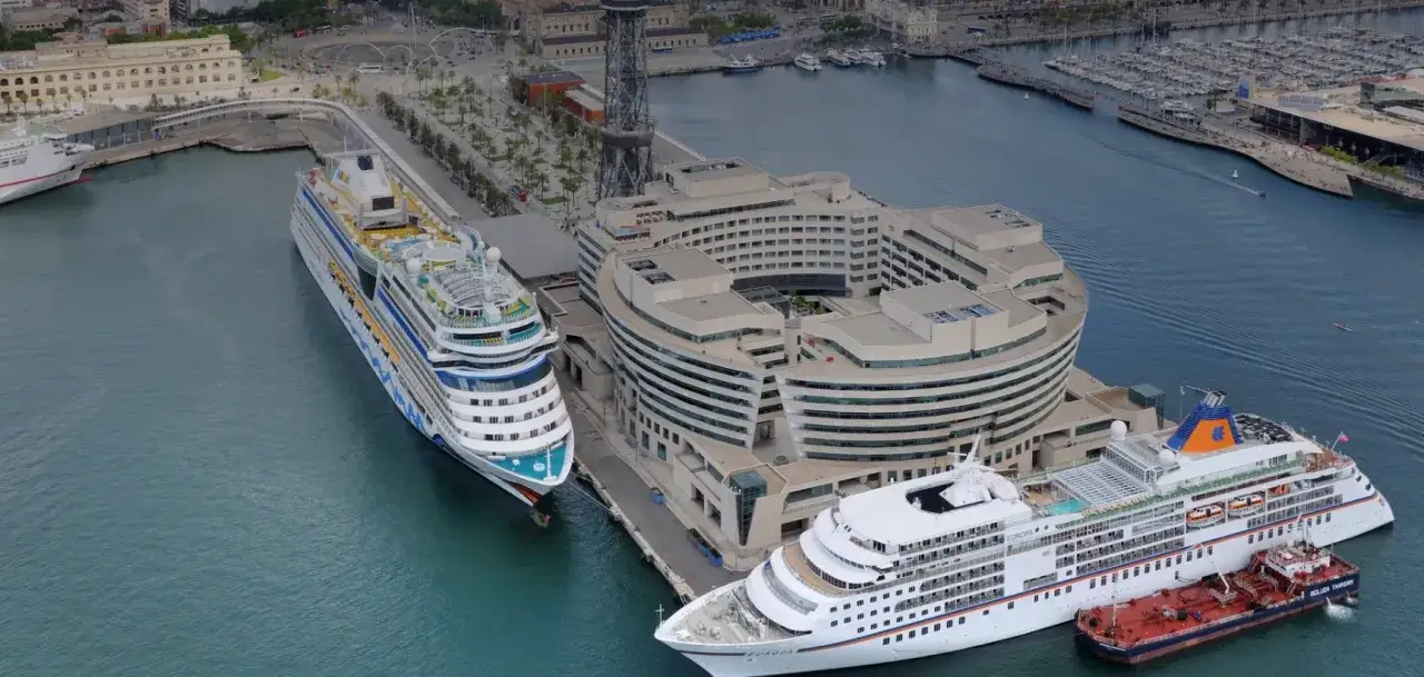 Grandi navi da crociera attraccate nel porto di Barcellona, vicino a un edificio moderno e a un porto turistico.