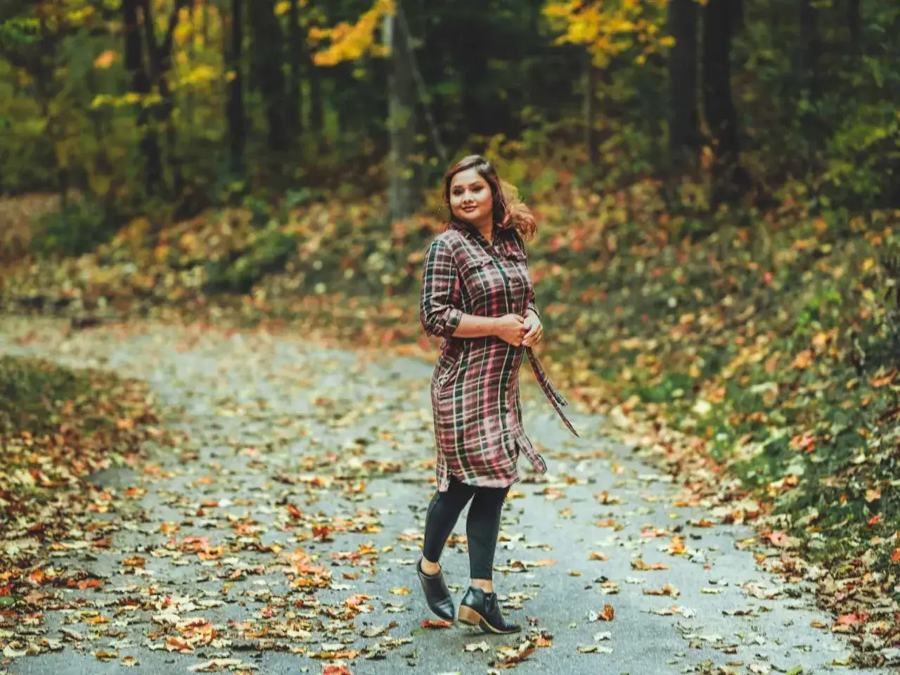 Frau im karierten Kleid mit Leggings kombiniert, steht auf einem herbstlich bedeckten Weg im Wald.