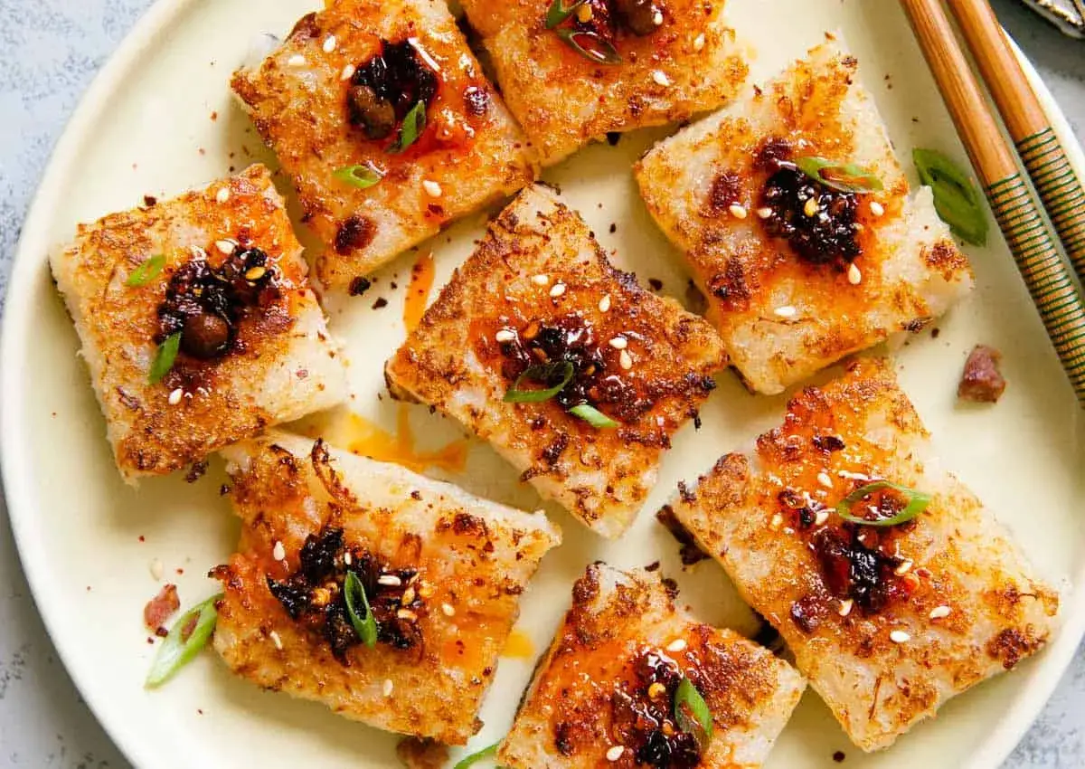 Golden-brown daikon mochi squares, pan-fried to perfection, are topped with a savory chili oil and sesame seed mixture, garnished with fresh scallions.