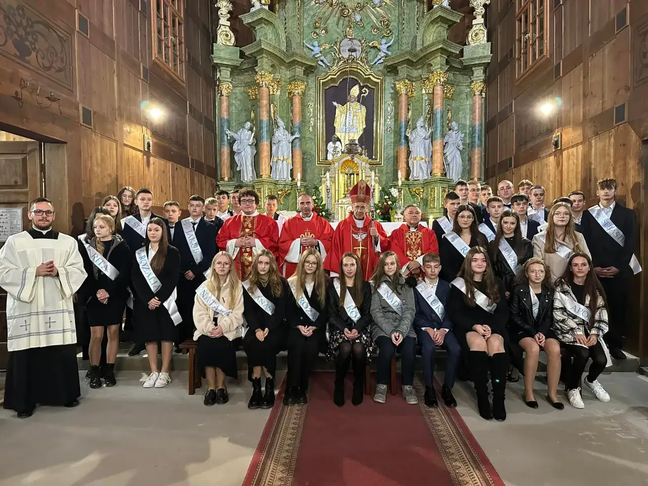 Grupa młodzieży w kościele, przygotowująca się do bierzmowania. W tle ołtarz, z przodu księża i bierzmowani w szarfach z imionami.