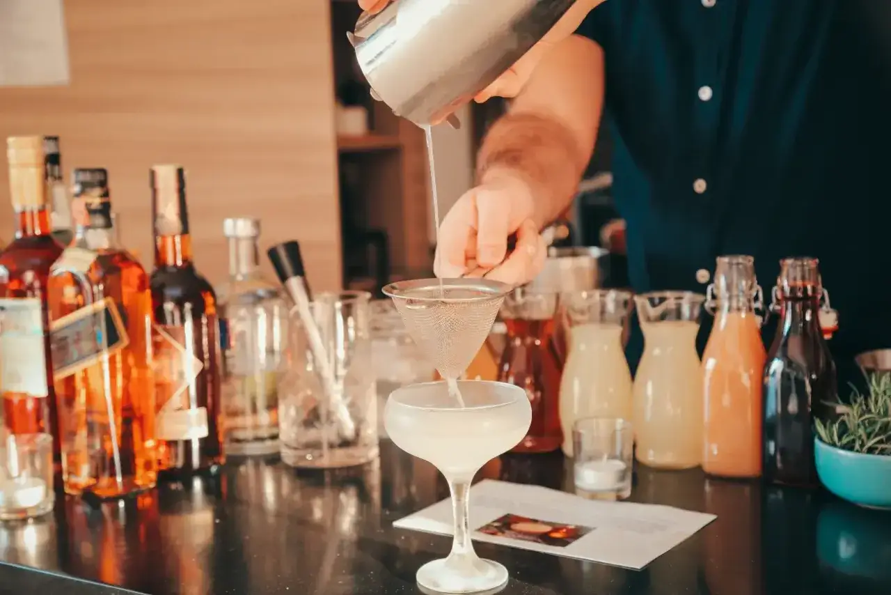 Barman przelewa drinka z shakera do drinków przez sitko do kieliszka. W tle butelki alkoholu i kolorowe syropy.