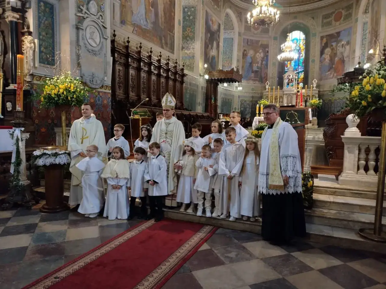 Dzieci w bieli po raz pierwszy przyjmują Komunię Świętą. Ceremonia w kościele trwa, a księża i rodzice towarzyszą im w tym ważnym dniu.