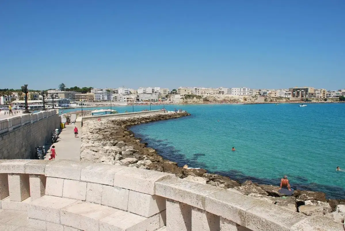 Un'incantevole giornata di sole a qui Salento, con il mare cristallino che lambisce la costa rocciosa e la citt&agrave; bianca sullo sfondo.