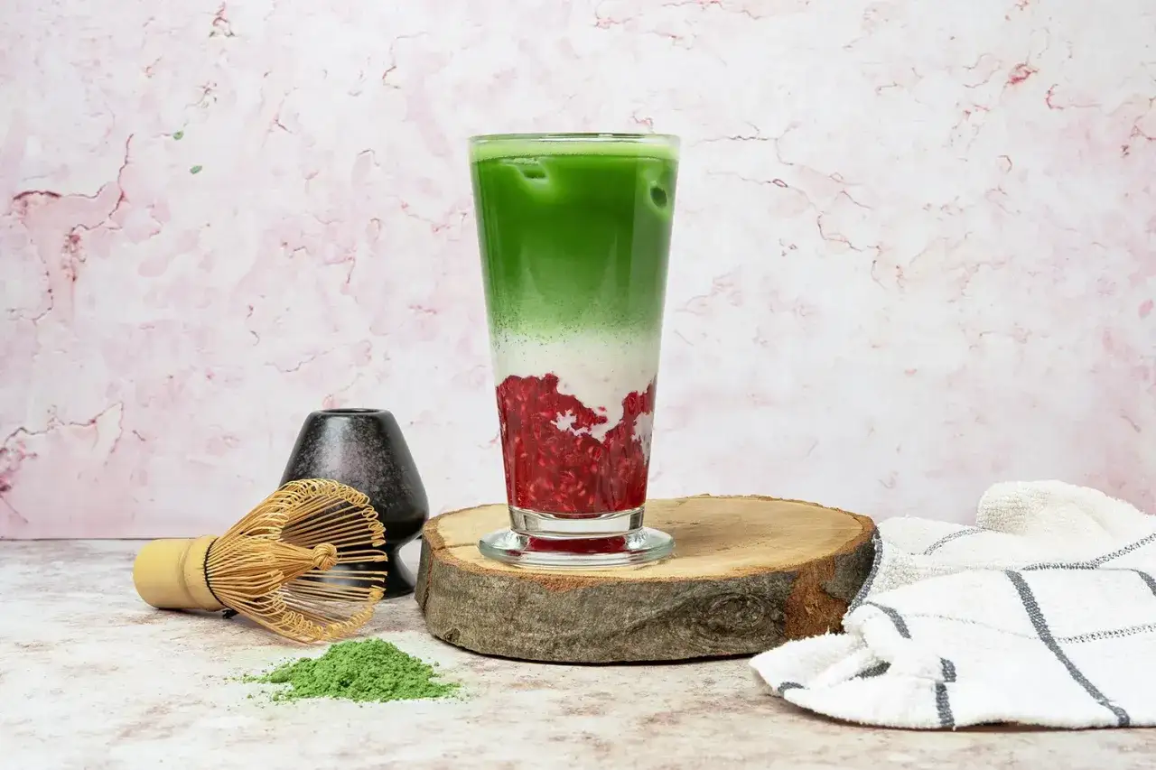 Orzeźwiające matcha z malinami w szklance, obok bambusowa miotełka i proszek matcha.