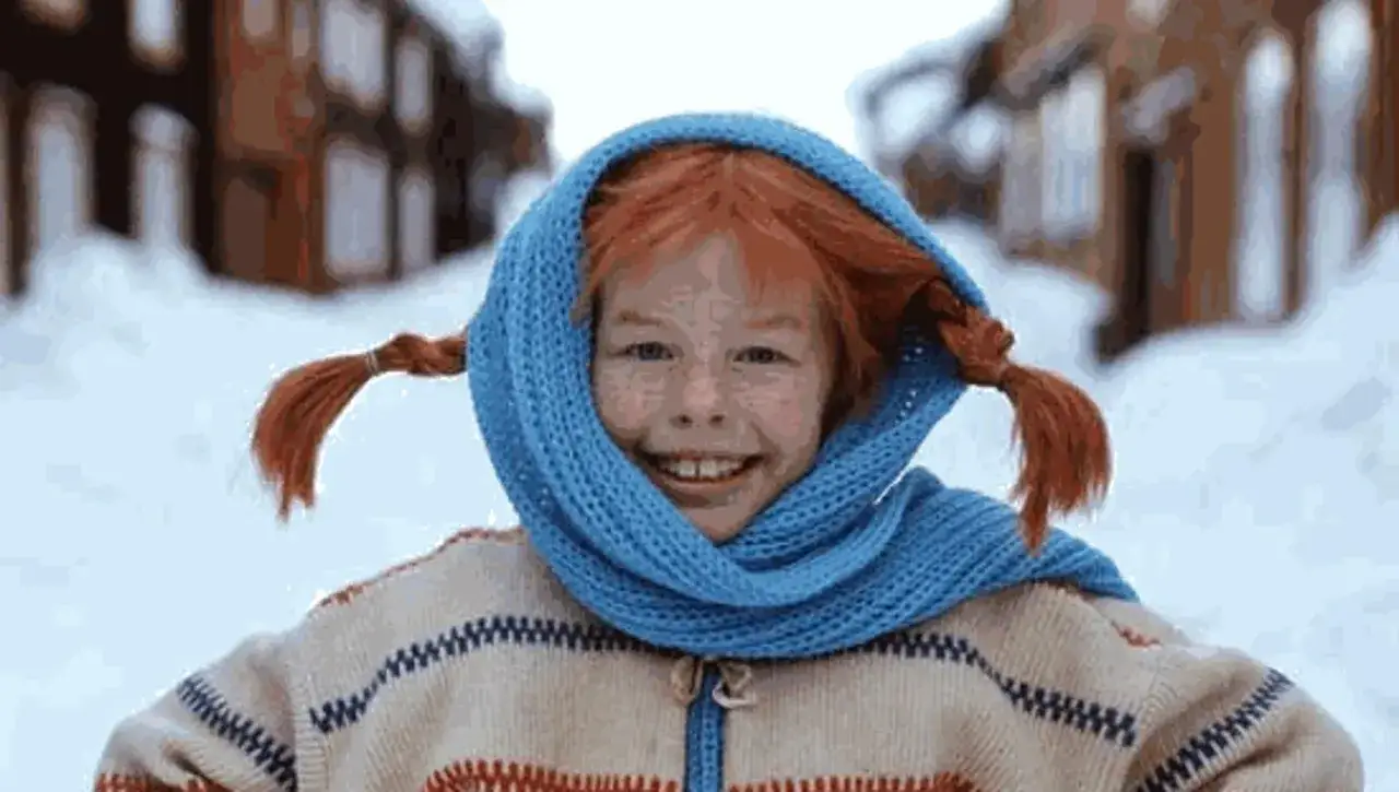 Pippi Calzelunghe, anche a 70 anni, sorride con le sue trecce rosse e un foulard blu, in un paesaggio innevato.