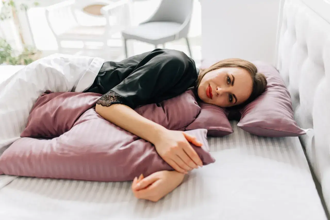 Donna in pigiama di seta nera, sdraiata su un letto con cuscini rosa, con evidenti rughe da cuscino sul viso.