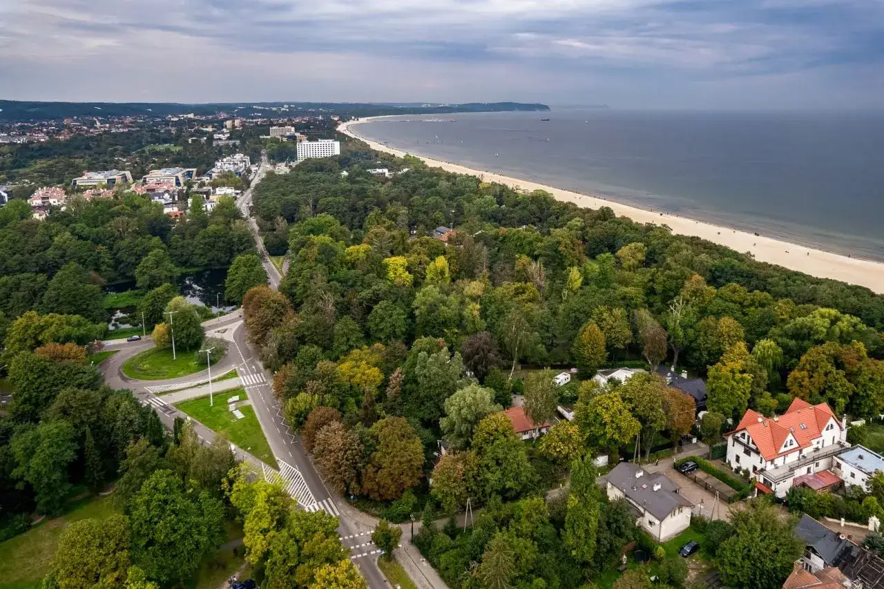Gdańsk Żabianka z lotu ptaka: zielone drzewa, plaża i morze. Widać zabudowania i drogi.