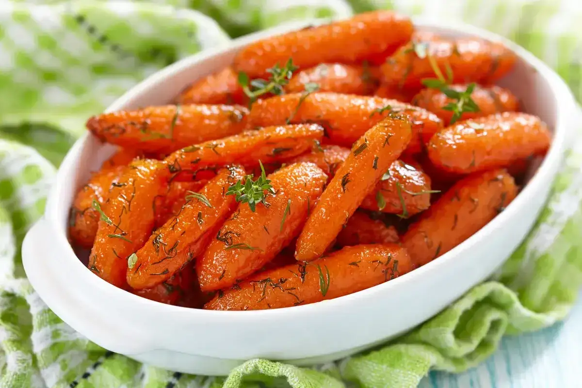 Carote glassate in agrodolce, guarnite con erbe aromatiche fresche, servite in una ciotola bianca su un tovagliolo verde.