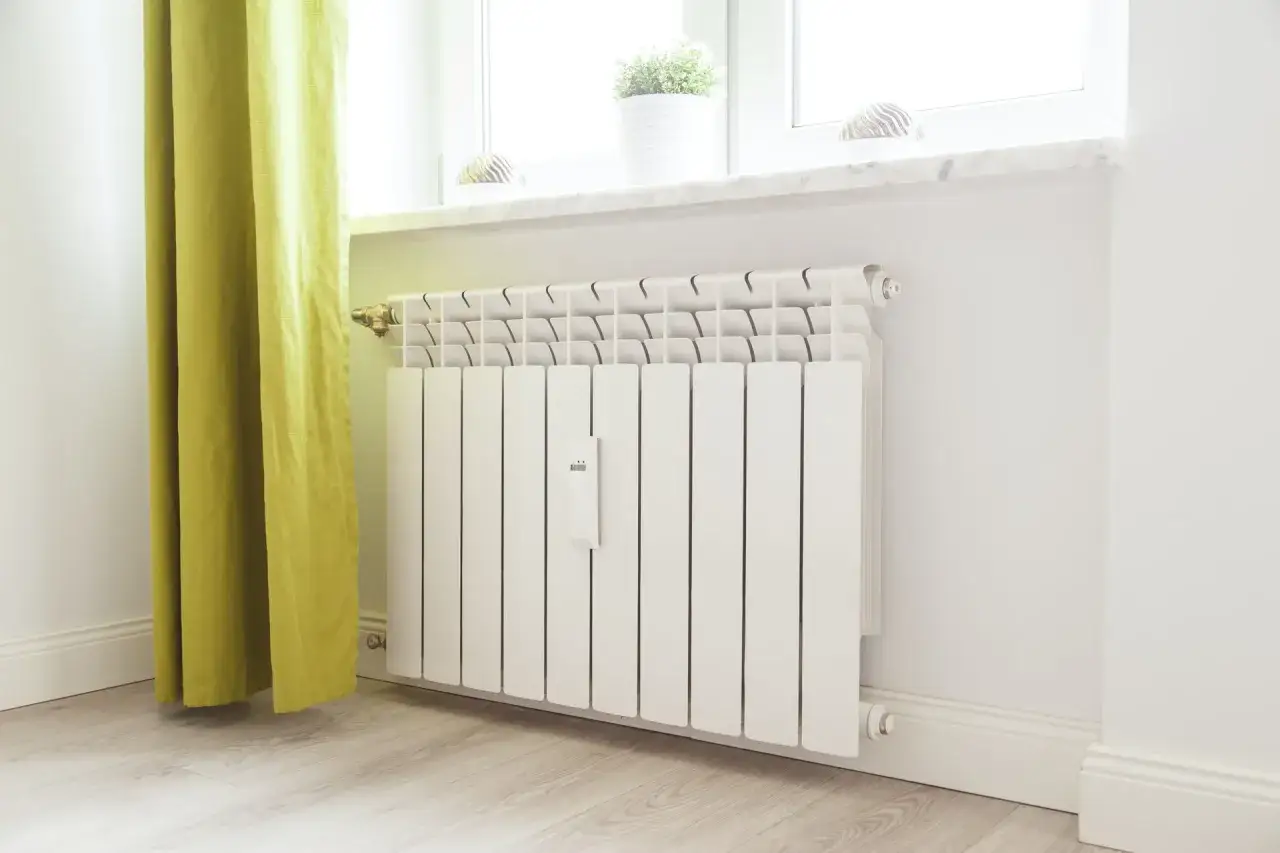 Biały grzejnik aluminiowy pod oknem, obok zielonej zasłony. Może być słychać szum w grzejnikach aluminiowych.