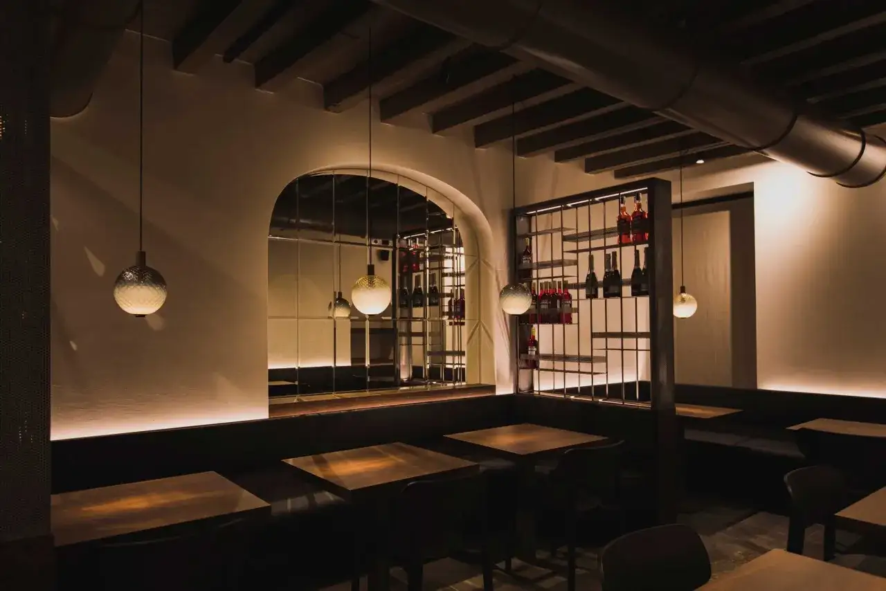 L'interno di un locale con tavoli in legno, sedute imbottite e un'ampia parete a specchio che riflette scaffali con bottiglie, evocando l'atmosfera de l'antico night club.