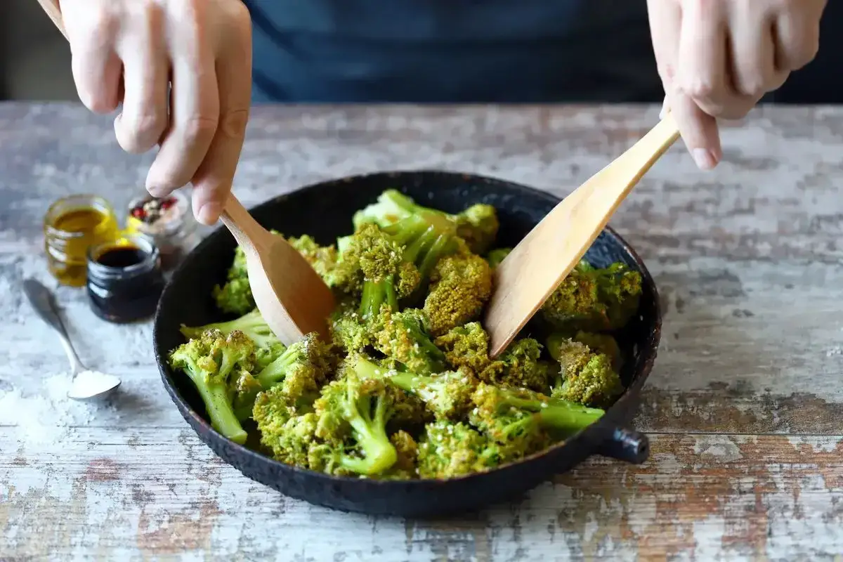 Mani che mescolano broccoli in padella, pronti per gustose ricette con broccoli. Olio, aceto e spezie a lato.