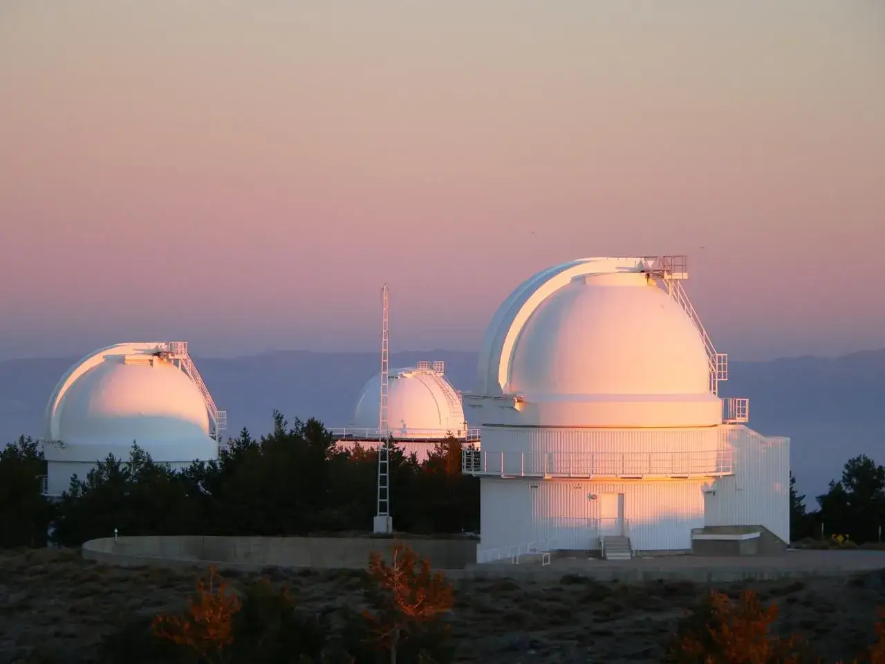Observatorios en el calar alto, con c&uacute;pulas blancas iluminadas por el atardecer, rodeados de &aacute;rboles y monta&ntilde;as al fondo.