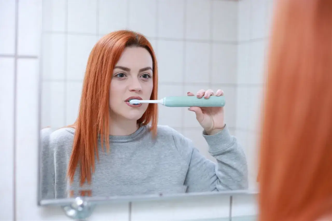 Ruda kobieta w szarej bluzie myje zęby szczoteczką soniczną, patrząc w lustro. To prosty sposób, jak myć zęby szczoteczką soniczną.