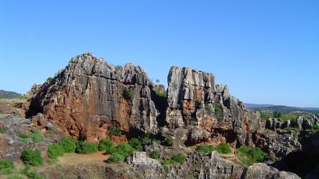 Majestuosas formaciones rocosas de tonos rojizos y grises se alzan bajo un cielo azul intenso. El **Cerro del Hierro** revela su geología imponente, salpicado de vegetación verde que contrasta con la piedra.