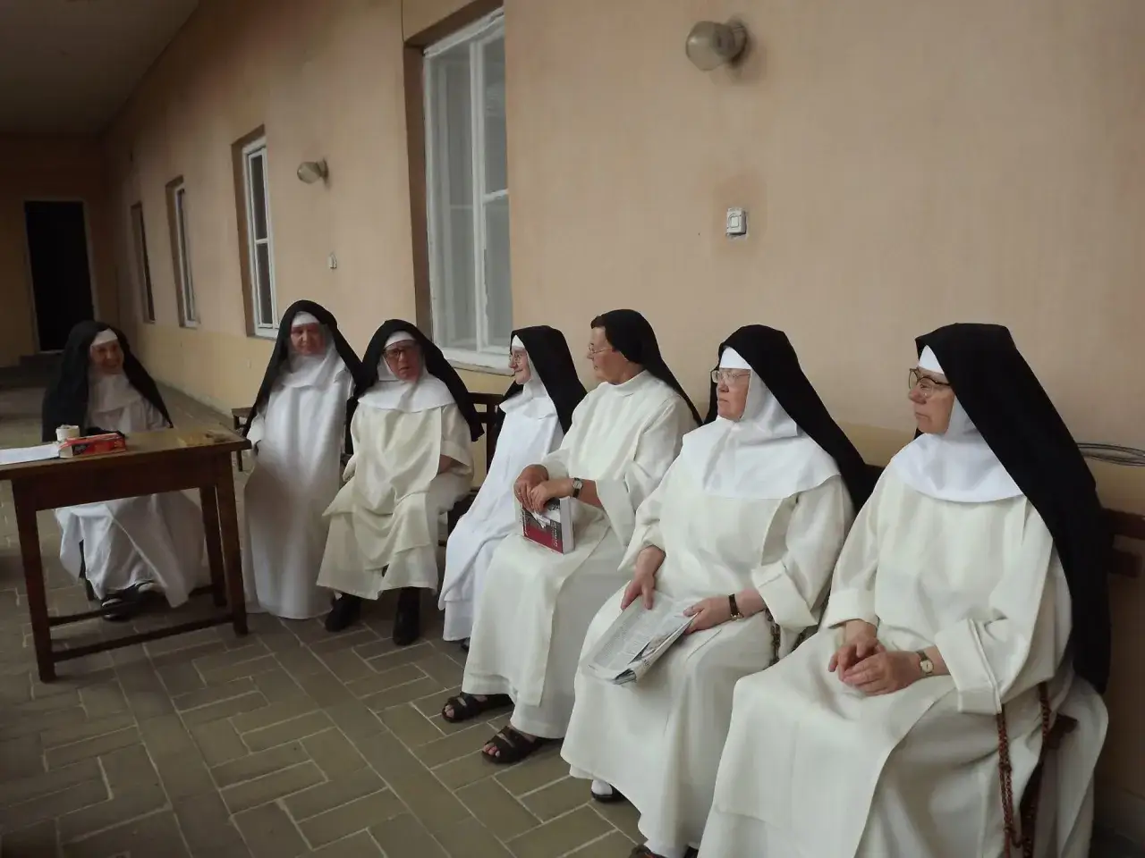 Siedem sióstr zakonnych w białych habitach i czarnych welonach siedzi w rzędzie. Jedna z nich uśmiecha się do aparatu.