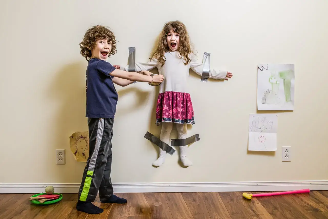 Ein Junge klebt seine Schwester mit Klebeband an die Wand. Sie sieht aus, als würde sie schreien. Dieser **kind macht alles kaputt**-Moment ist ein Beweis für ihre verspielte Natur.