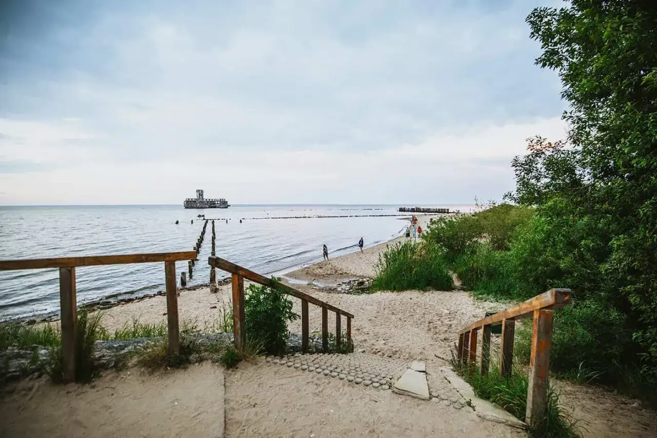 Piękne, tanie domki letniskowe nad morzem czekają! Zejście na plażę z drewnianymi schodkami, morze i tajemnicza budowla na horyzoncie.