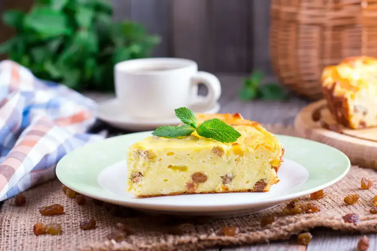 Una fetta di torta putana, soffice e dorata, con uvetta e una fogliolina di menta, accanto a una tazza di caffè.