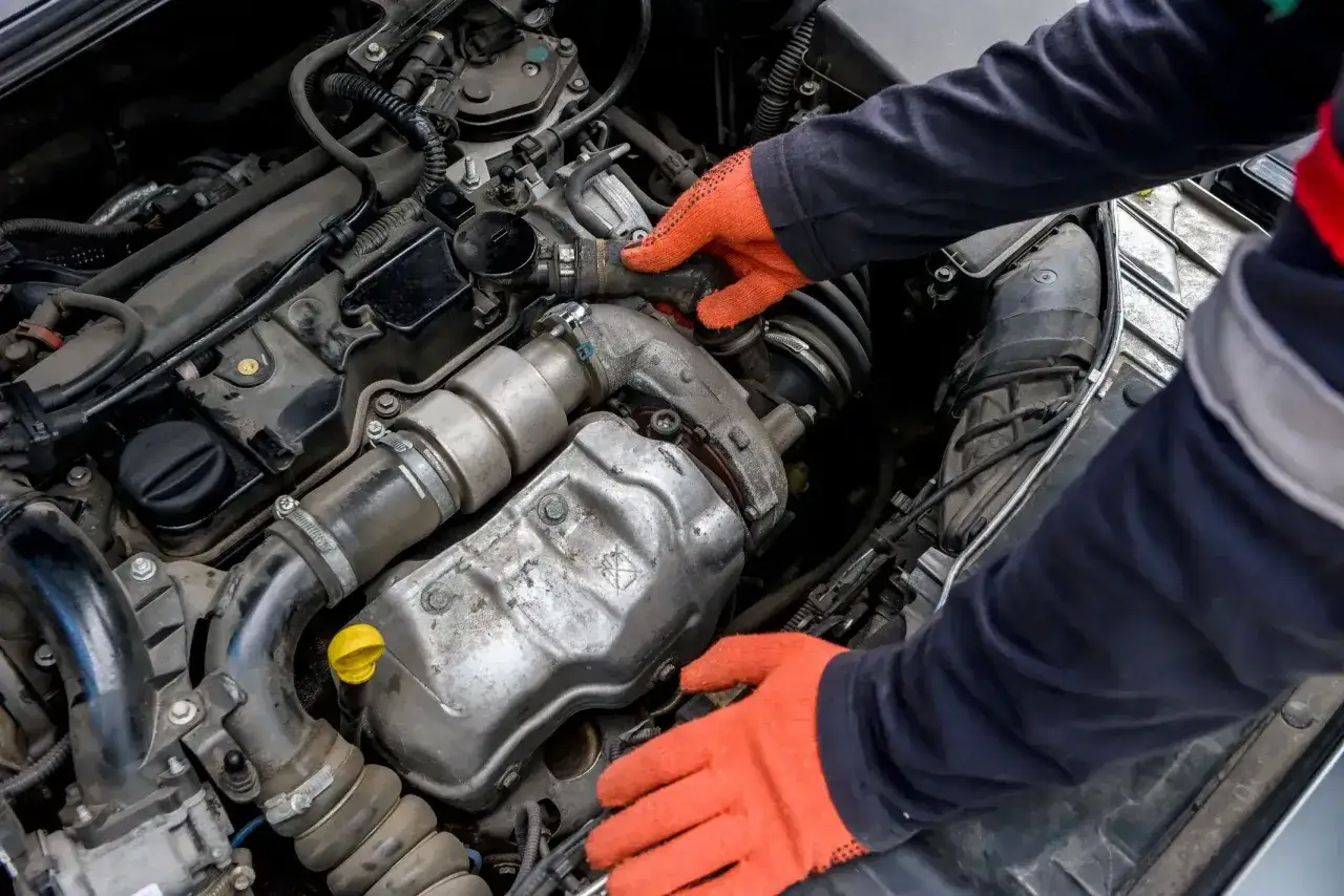 Mechanik w pomarańczowych rękawiczkach sprawdza silnik, szukając przyczyny stukania na zimnym silniku.