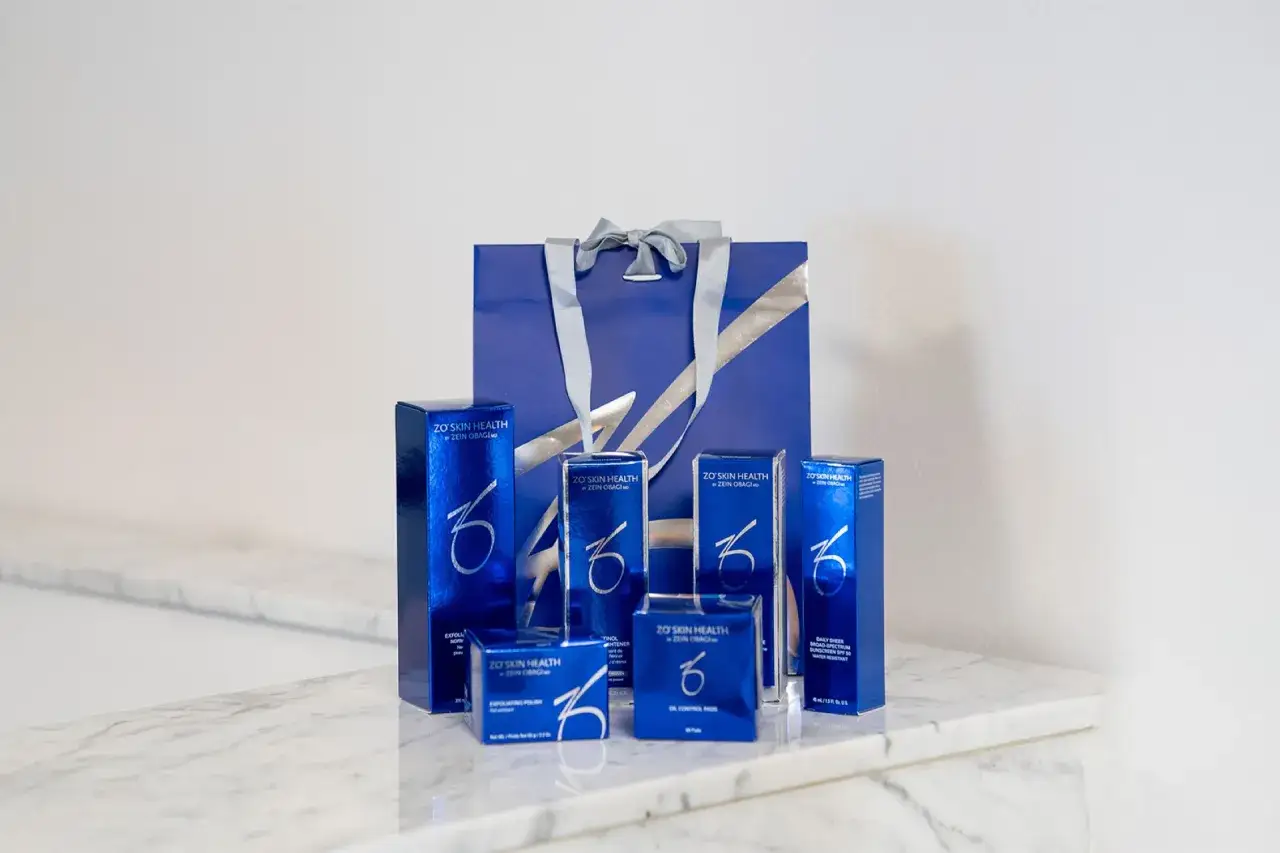 En samling ZO Skin Health-produkter, skapade av Zein Obagi, står uppradade framför en blå presentpåse.