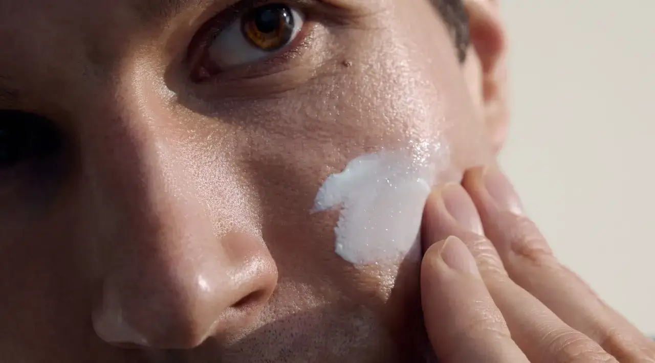Primo piano di un uomo che applica crema sul viso. La sua pelle &egrave; luminosa, segno di una perfetta pulizia viso uomo.
