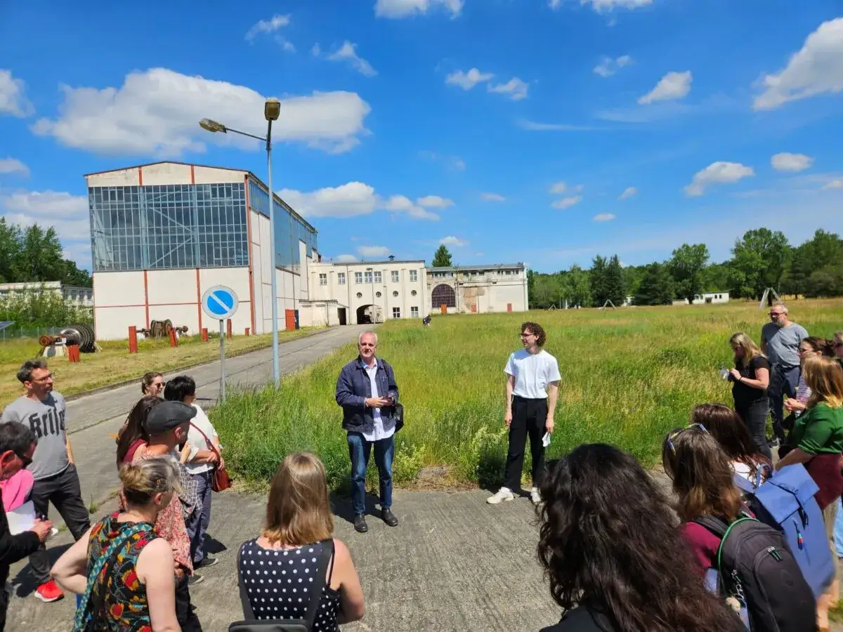 Gruppe bei einem **Festival im Osten** vor Industriegeb&auml;uden. Ein Mann erkl&auml;rt etwas, w&auml;hrend andere zuh&ouml;ren.