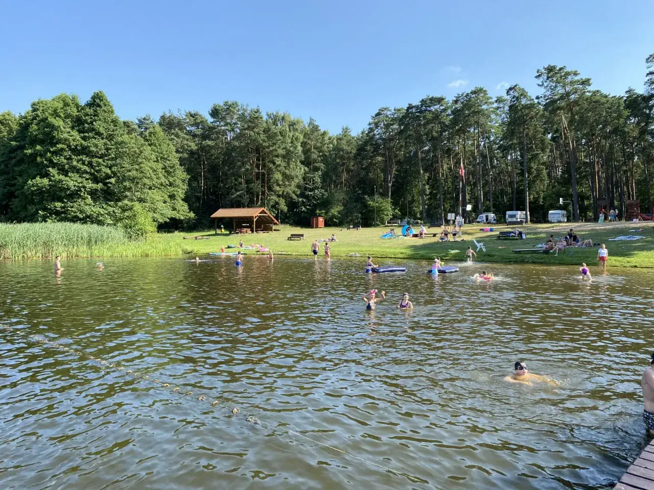 Słoneczny dzień nad jeziorem Szczutowo. Ludzie kąpią się, opalają na trawie i odpoczywają pod wiatą.