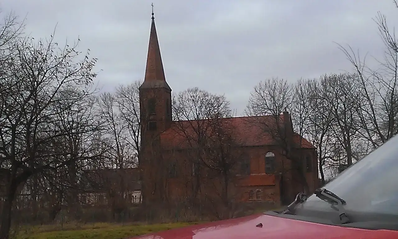 Gotycka ceglana świątynia w Duszniki Wielkopolskie, otoczona nagimi drzewami, z czerwonym dachem i strzelistą wieżą.
