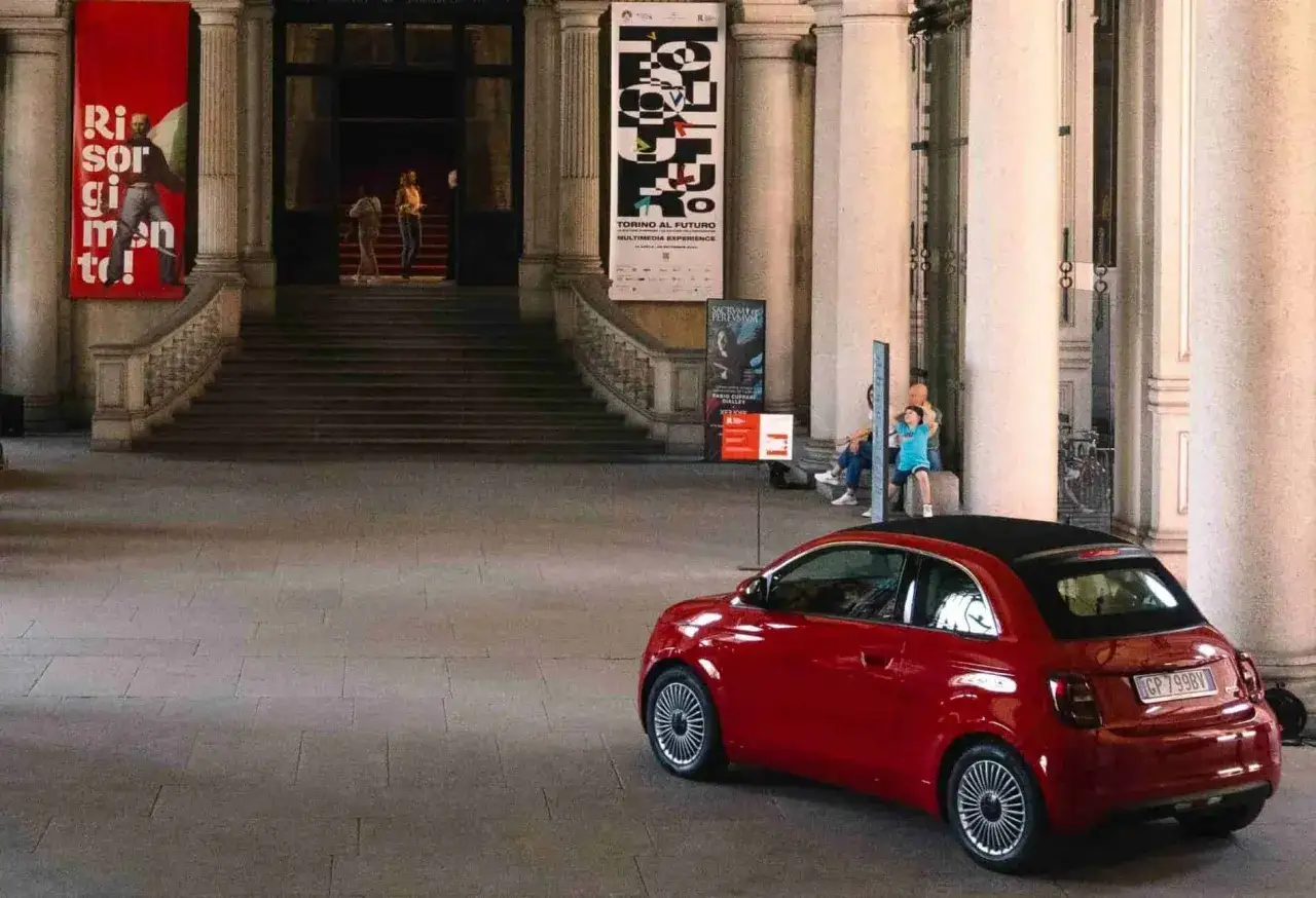 Czerwony fiat 500, znany z Włoch, parkuje przed budynkiem z banerami.