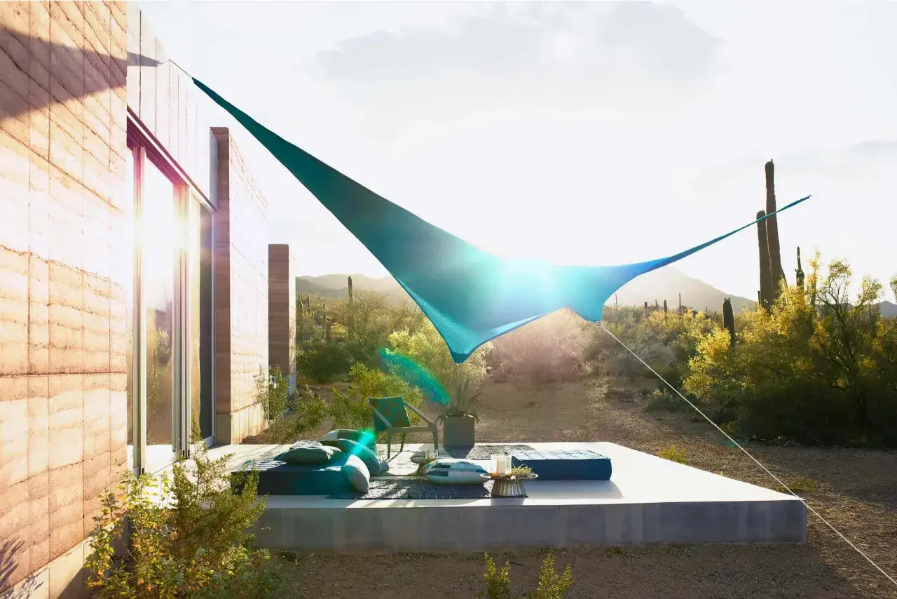 Ideas para dar sombra en un patio: una vela azul tensada sobre un &aacute;rea de descanso con cojines y un sill&oacute;n, rodeada de cactus y vegetaci&oacute;n des&eacute;rtica.