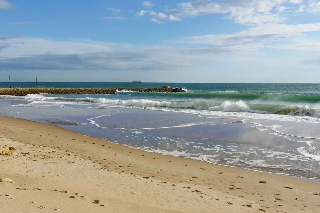 Strand von Rota, Spanien. Wellen brechen an einem steinernen Wellenbrecher, w&auml;hrend ein Schiff am Horizont vorbeif&auml;hrt.