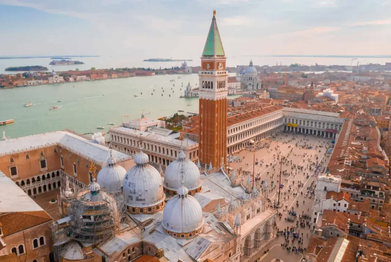 Vue aérienne de la **Piazza San Marco** à Venise, avec la basilique, le campanile et le canal. Une foule anime la place.