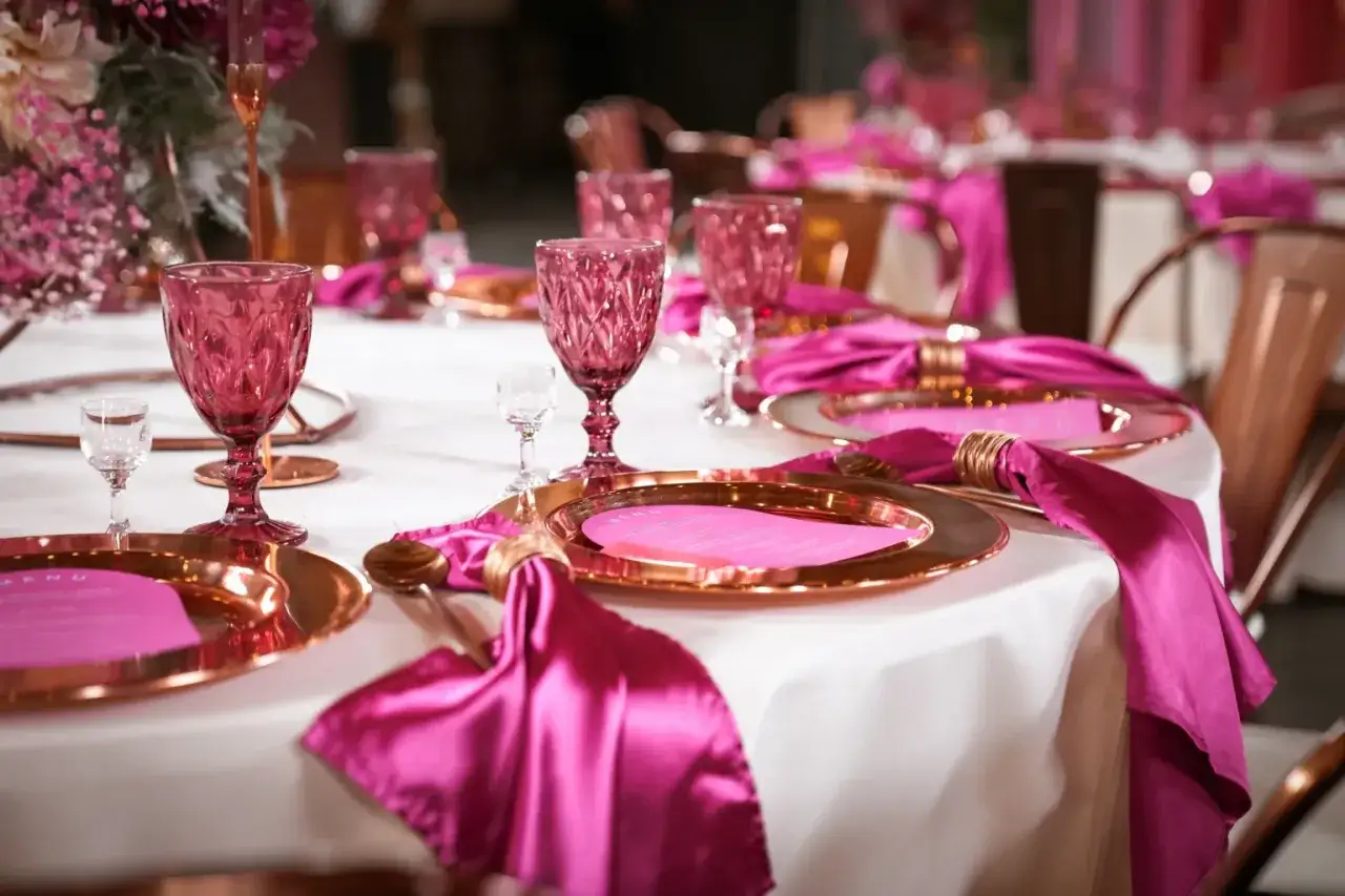 Elegancko ułożone, fuksjowe serwetki na stole, obok złotych talerzy i różowych kieliszków, tworzą zachwycającą aranżację.